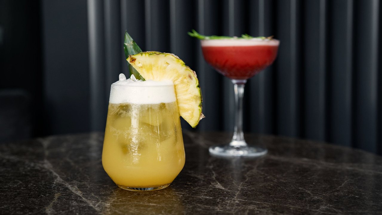 Fotografía de referencia: dos cocteles, uno con piña y otro rojo en copa, sobre una mesa de mármol oscuro.
