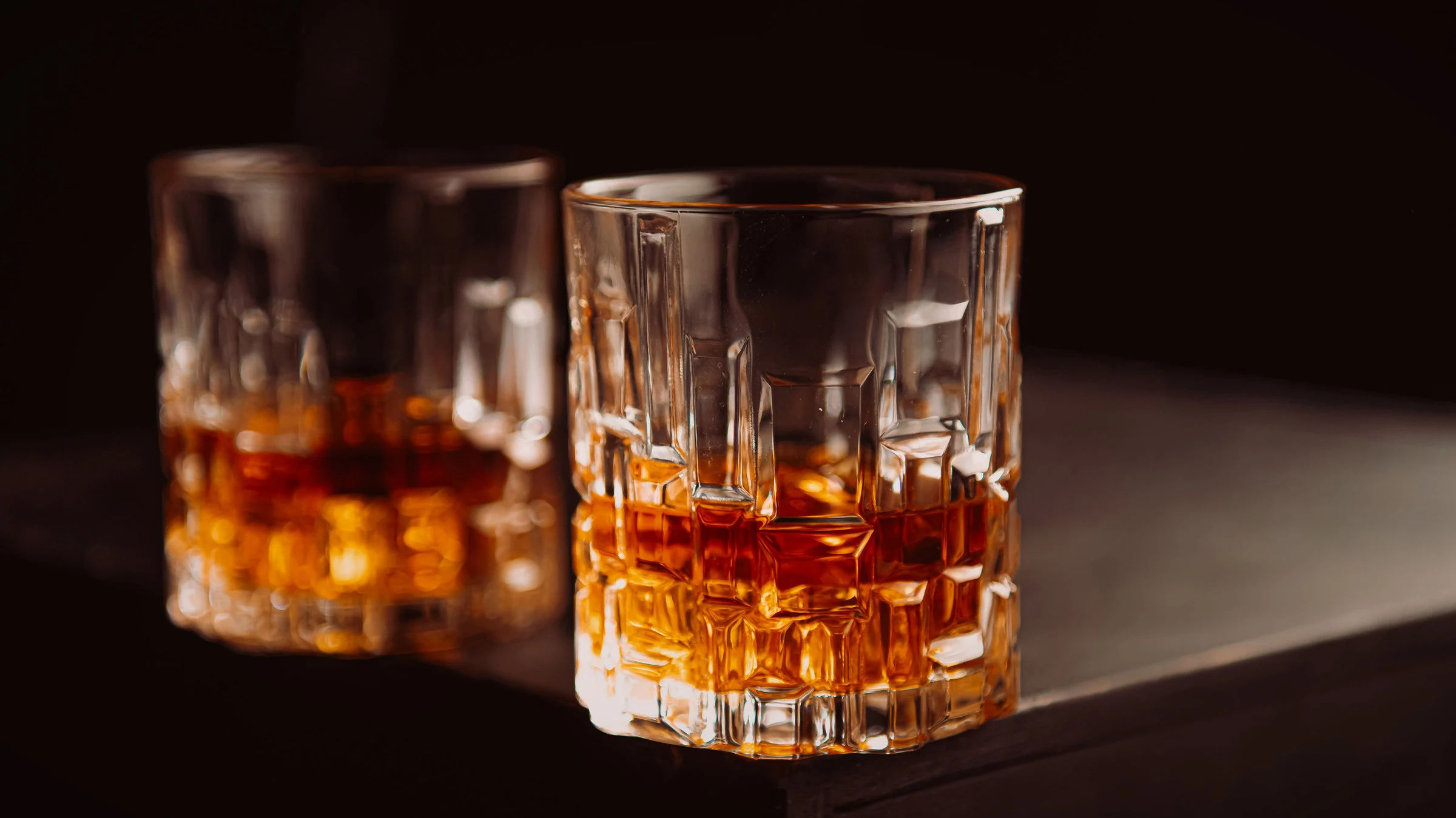 Fotografía de referencia: dos vasos de cristal cortado con whisky sobre una barra oscura, sin marcas visibles.