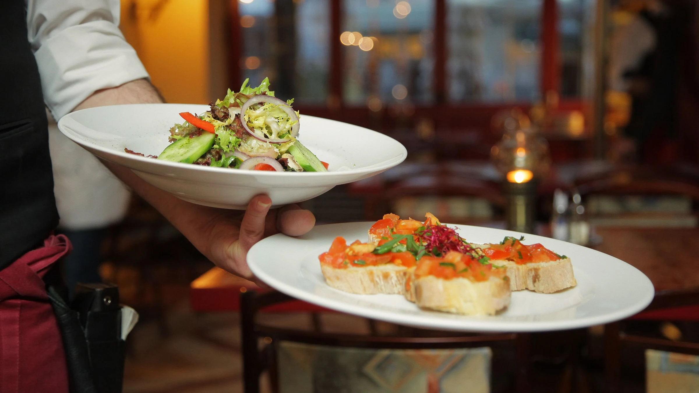 Fotografía de referencia: un mesero lleva una ensalada y unas tostadas a la mesa en un restaurante cálido.