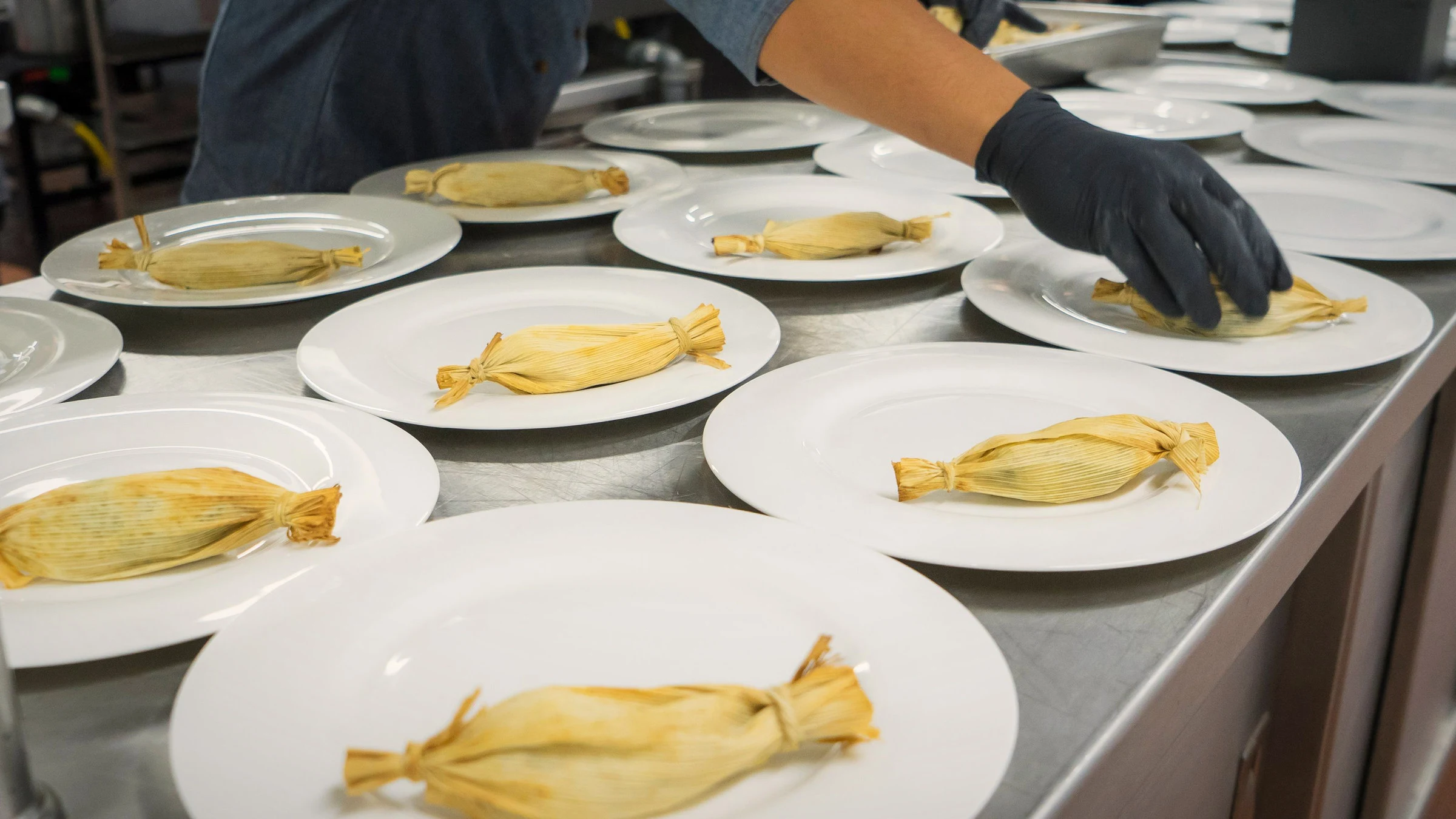 Fotografía de referencia: una mano con guante negro emplata tamales iguales sobre una mesa de acero.