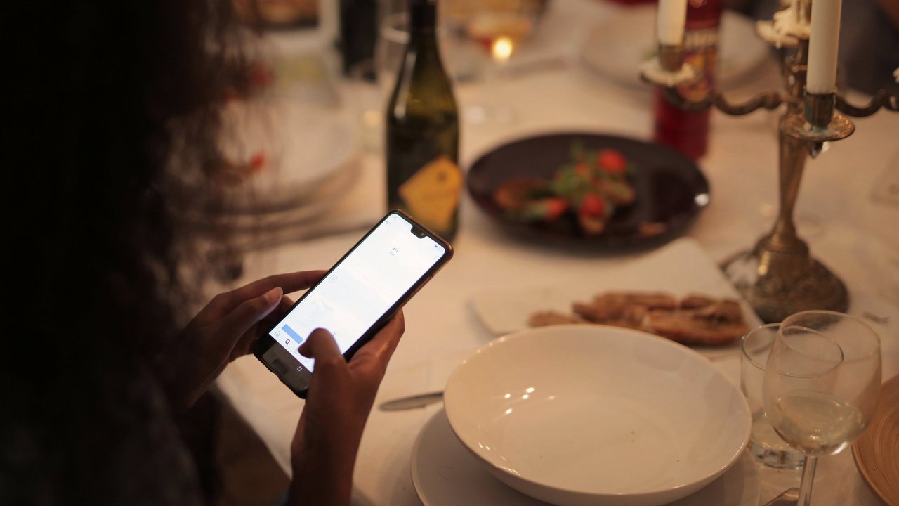 Fotografía de referencia: una persona consulta su celular en una mesa servida a la luz de las velas.