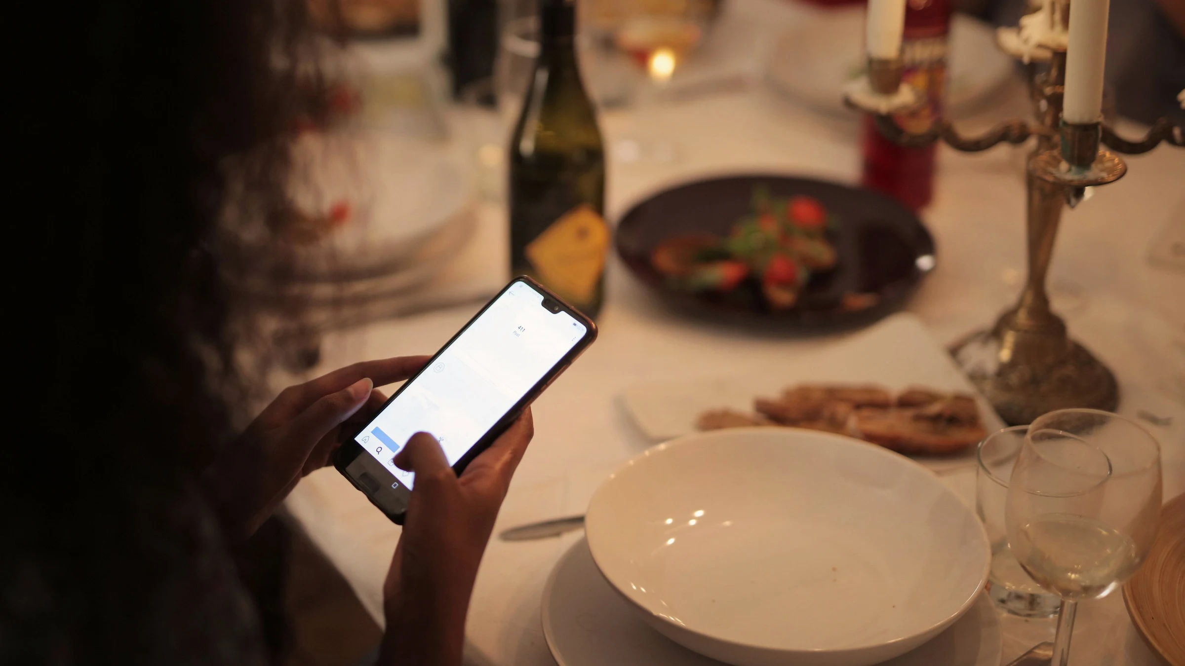 Fotografía de referencia: una persona consulta su celular en una mesa servida a la luz de las velas.