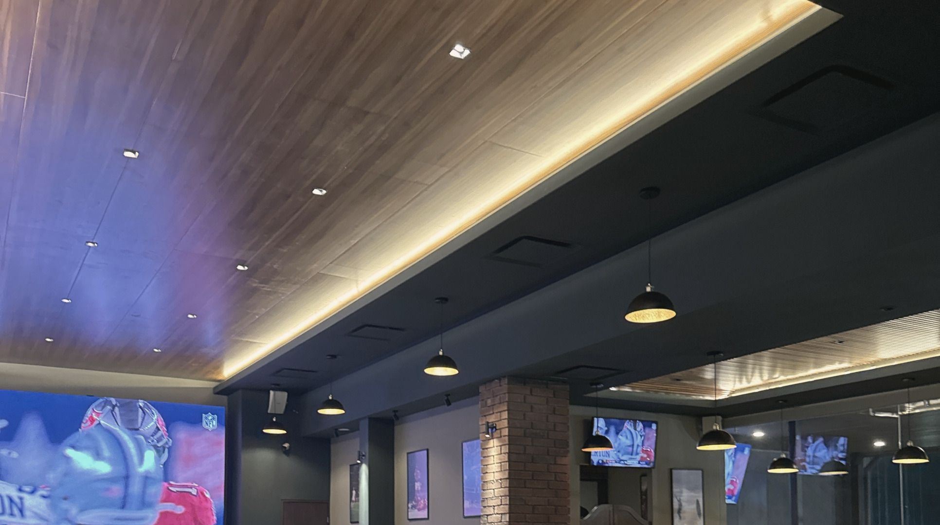 Sala del Bar La Cumbre con techo de madera iluminado, lámparas colgantes, una columna de ladrillo y pantallas con un partido.