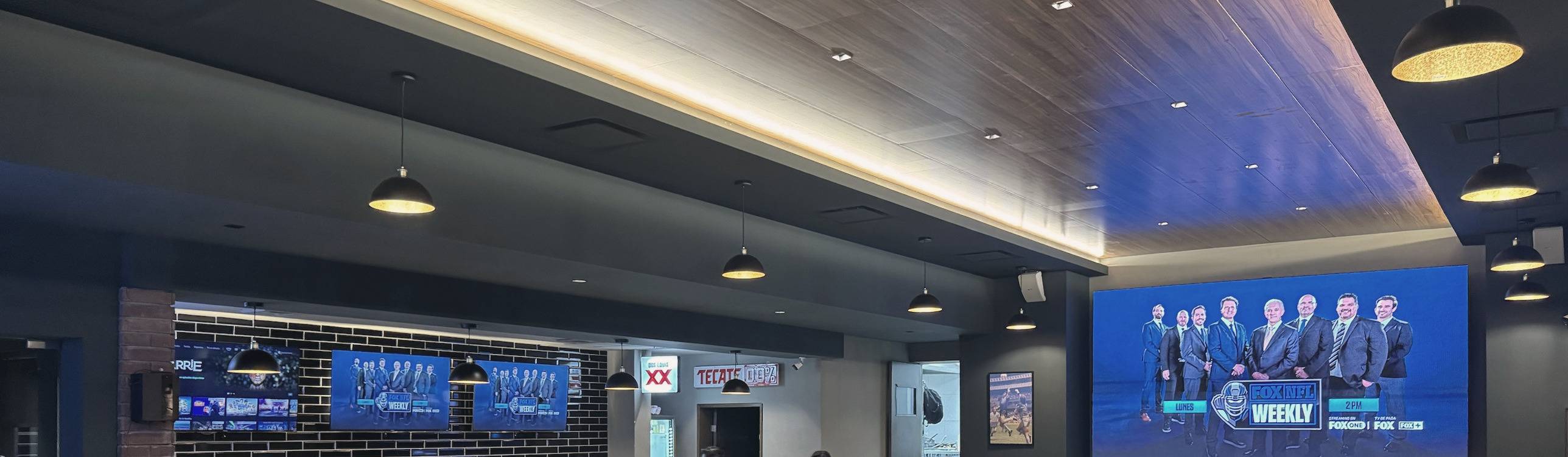 Vista panorámica del Bar La Cumbre con techo de madera iluminado, lámparas colgantes, la barra con televisiones y una pantalla gigante.