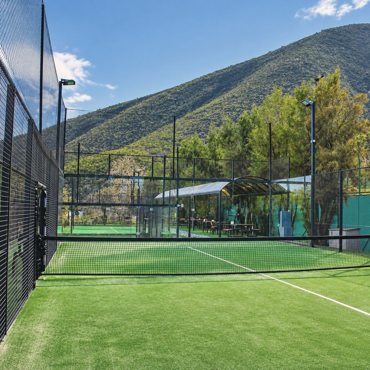 Cancha de pádel de pasto sintético verde con muros de cristal y malla, con el cerro arbolado al fondo.
