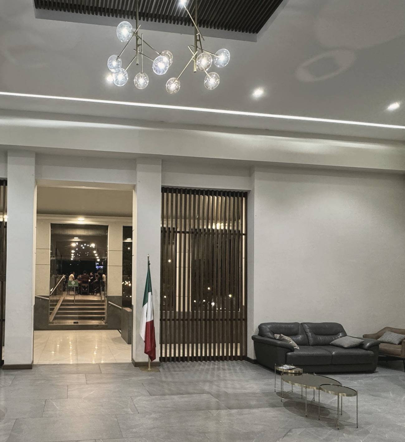 Vestíbulo del club con piso de piedra gris, lámpara de esferas de cristal, celosías de madera, una sala de piel y la bandera de México junto al paso hacia el bar.
