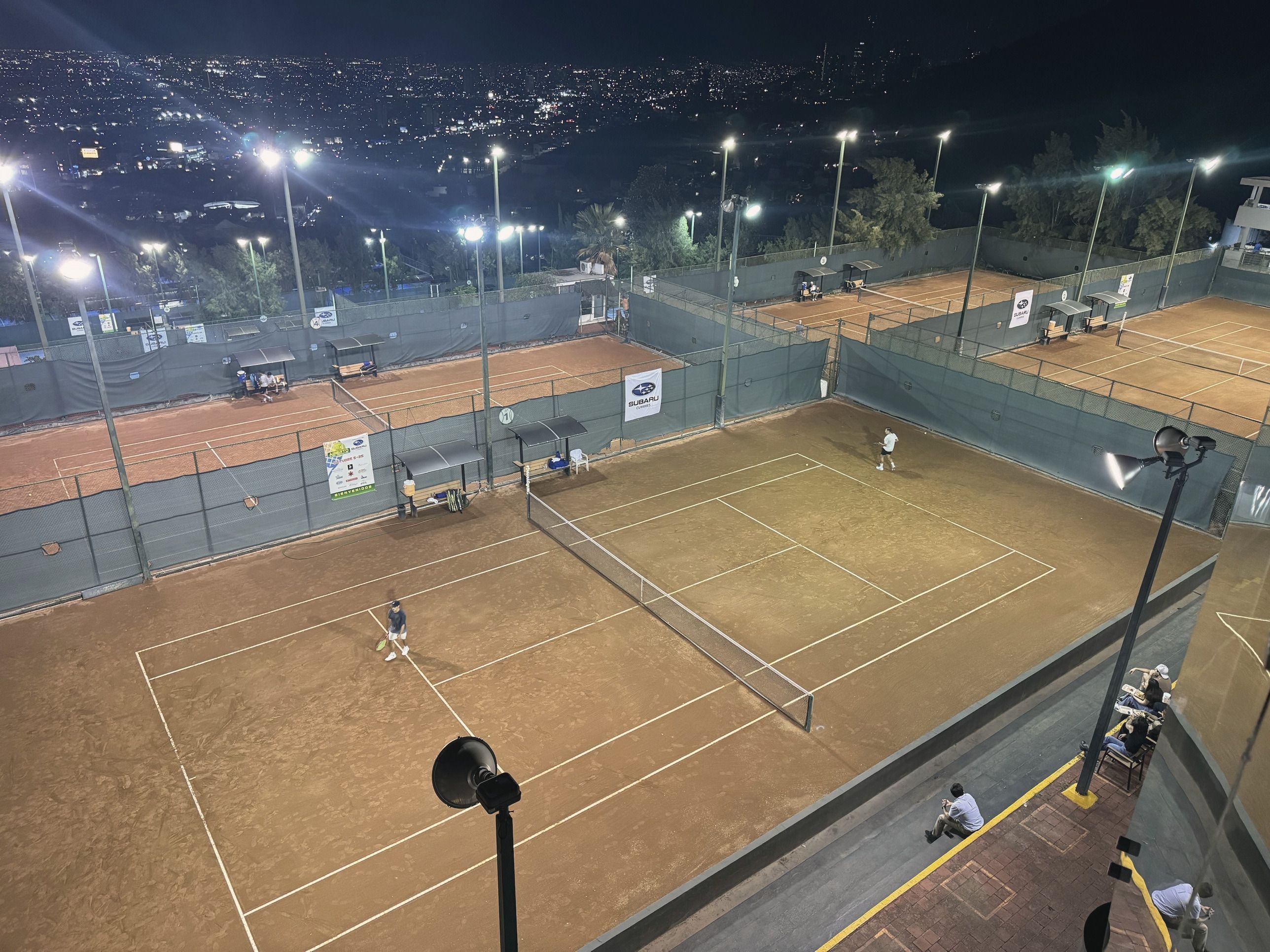 Canchas de tenis de arcilla iluminadas de noche, vistas desde lo alto, con las luces de Monterrey al fondo.